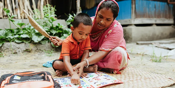 A woman sits on a woven mat with a young child, pointing to pictures in a colorful learning book outdoors