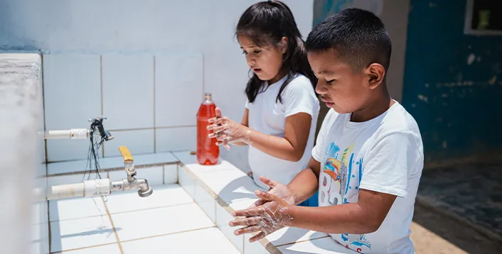 Two children wash their hands with soap at an outdoor sink.